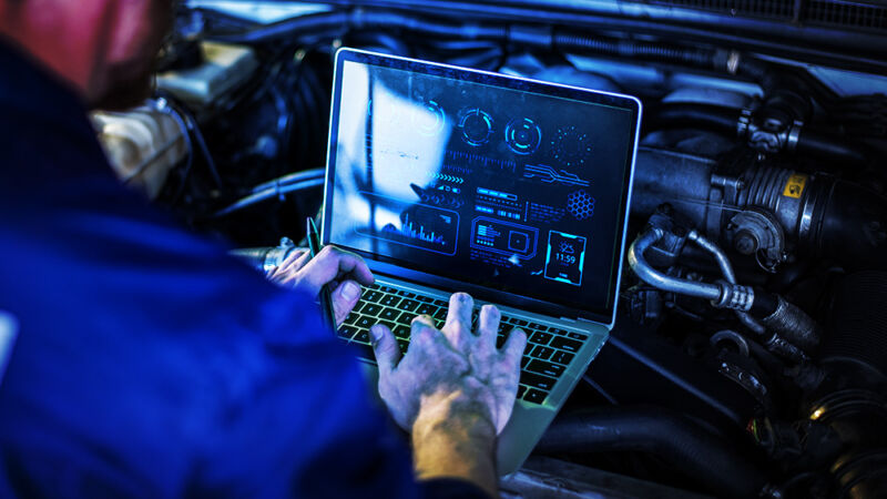 A man operates a notebook computer over the open engine of a car.